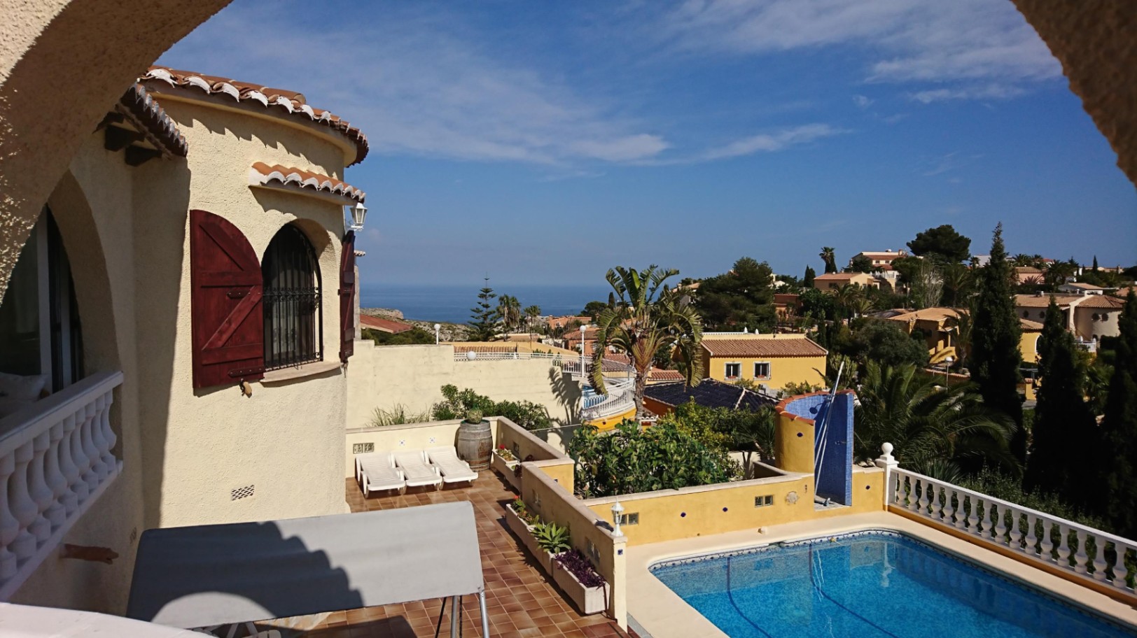 Exterior view featuring a rectangular pool, terracotta sun terrace with loungers, and a villa tower with wooden shutters overlooking the sea.