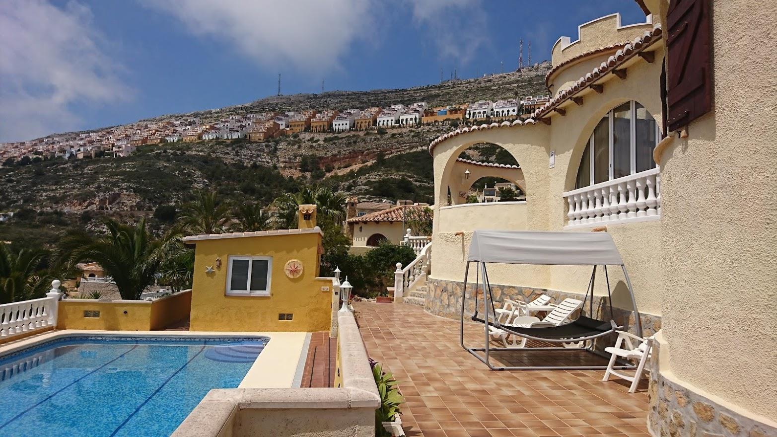 Exterior view featuring the swimming pool, tiled terrace with a sun swing, and arched naya. The hillside urbanization of Cumbre del Sol is visible in the background.