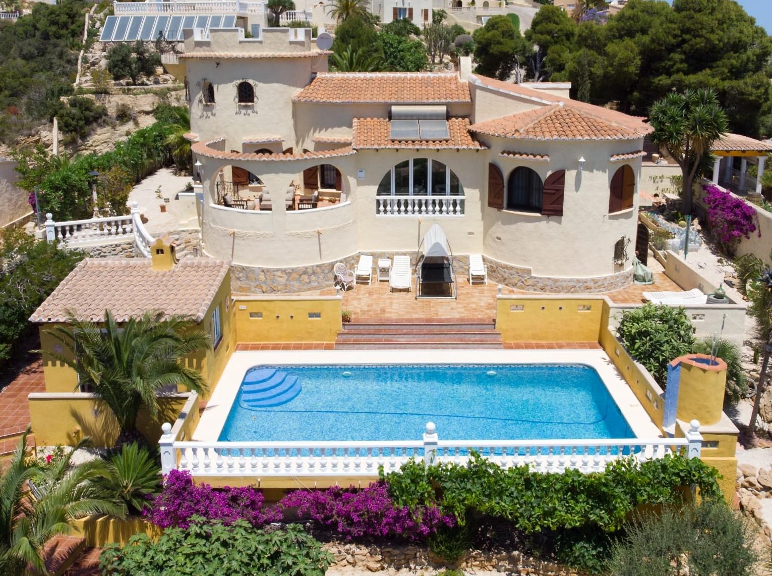 Exterior view of the villa featuring a rectangular pool, traditional tower architecture, terracotta roofs, and a landscaped garden with Mediterranean flora.