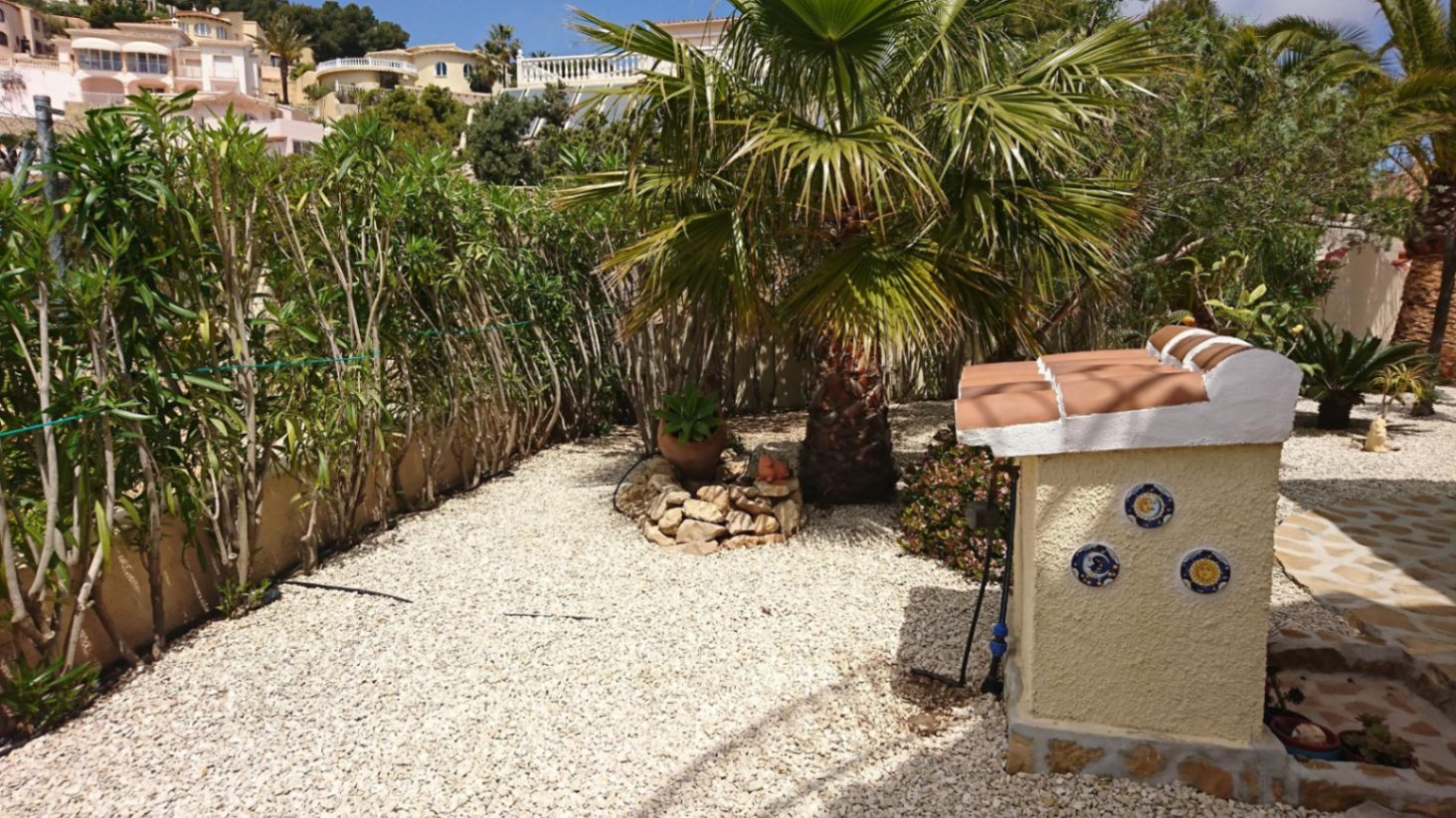 Mediterranean garden featuring white gravel landscaping, a mature palm tree, and a decorative stone pedestal with traditional ceramic tiles.