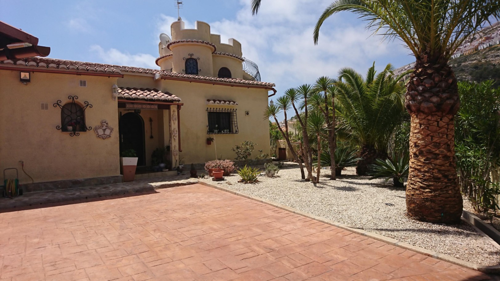 Mediterranean villa exterior featuring a crenelated tower, terracotta tiled driveway, and a low-maintenance gravel garden with mature palm trees.