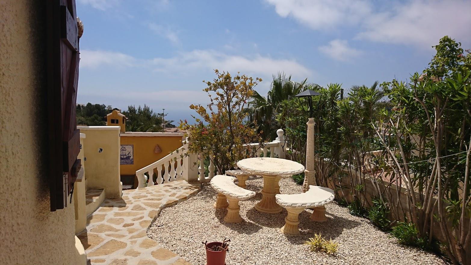 Outdoor terrace featuring a carved stone table and benches on a gravel surface, bordered by Mediterranean plants and a white balustrade with sea views.