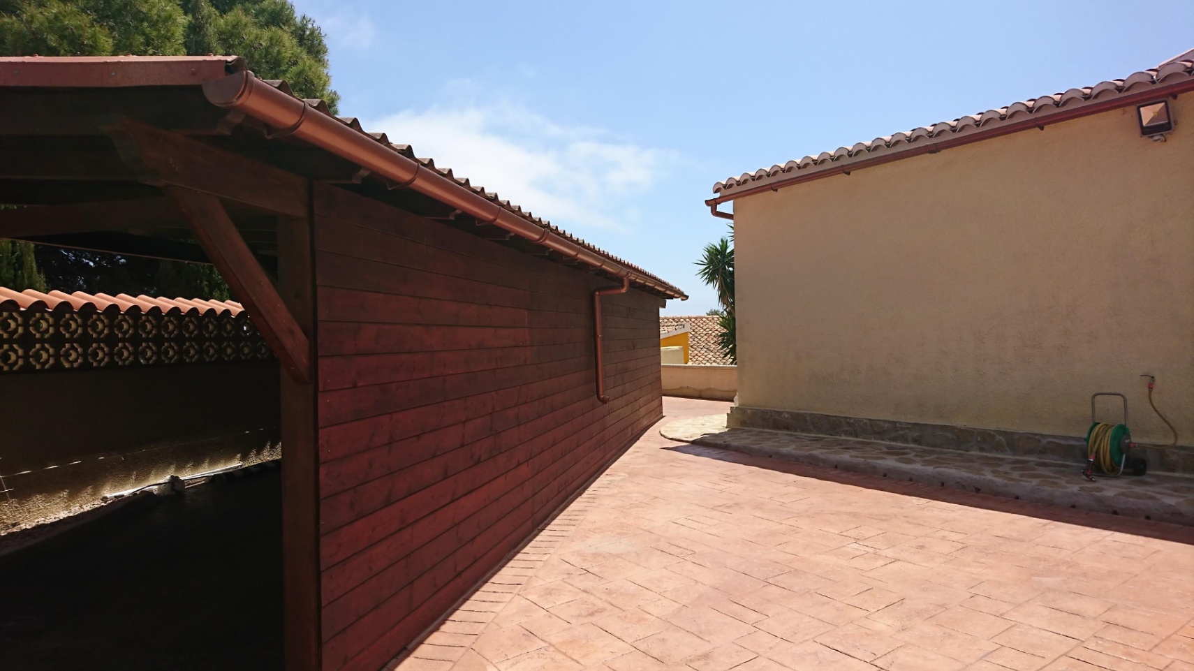 Paved outdoor area featuring a wooden carport or storage structure alongside the main building's facade, both finished with traditional ceramic tile roofs.