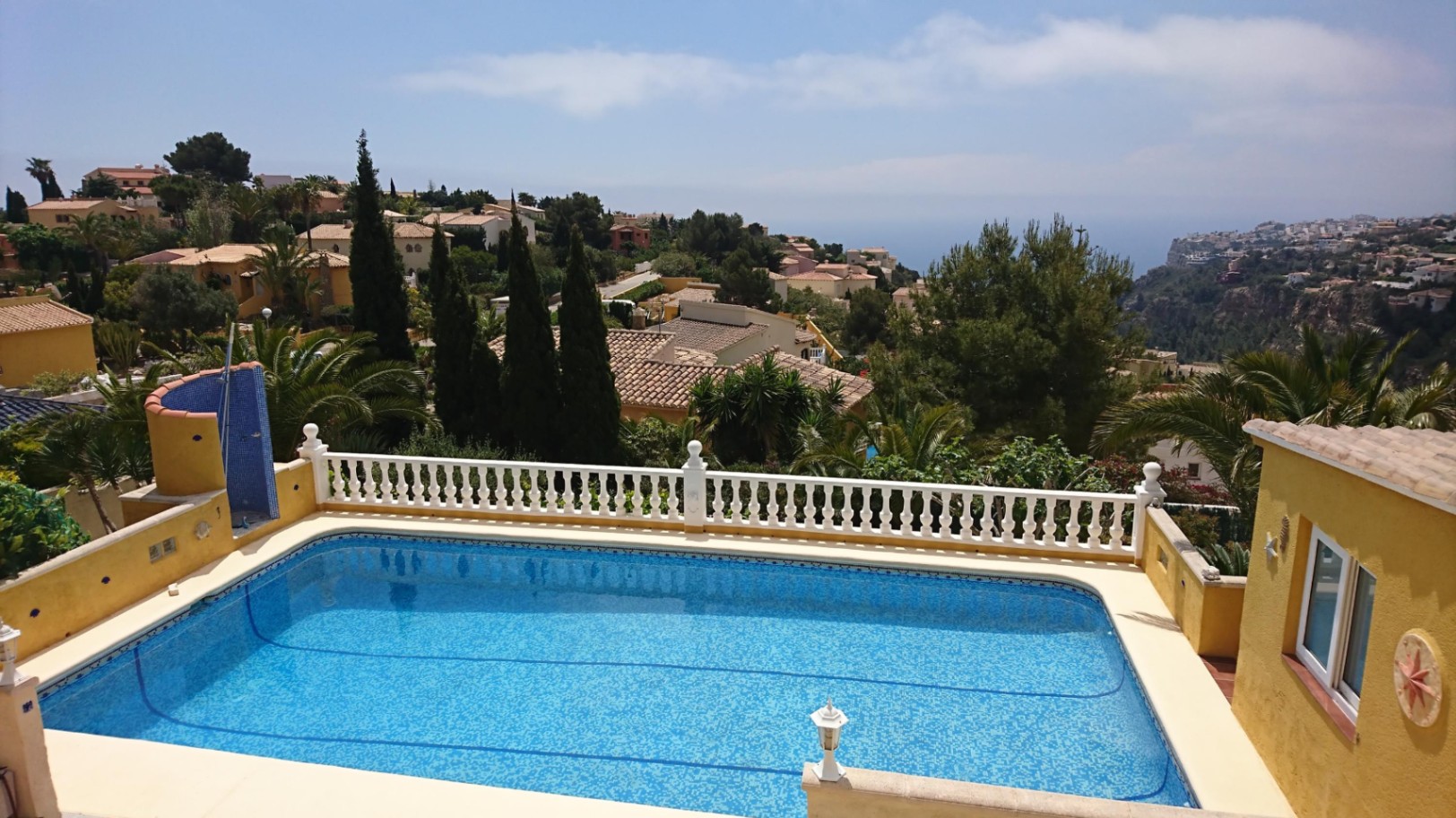 Rectangular swimming pool with blue mosaic tiling, featuring a classic white balustrade and panoramic views of the Mediterranean coastline and surrounding greenery.