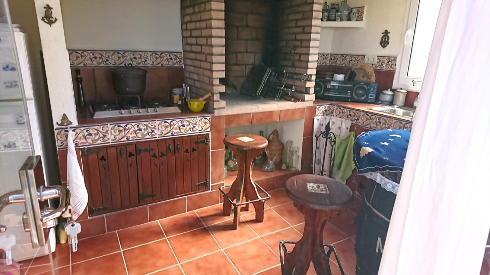 Rustic summer kitchen featuring a built-in brick barbecue, decorative tile splashbacks, wooden cabinetry, and traditional terracotta flooring.