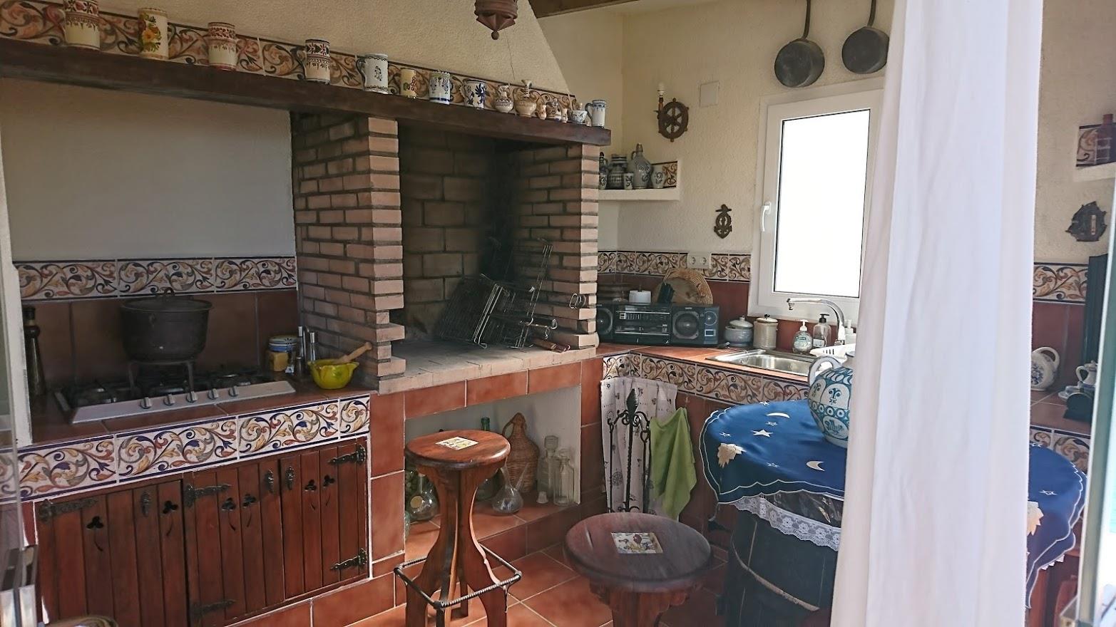 Rustic summer kitchen featuring a built-in brick barbecue, traditional patterned tilework, wooden cabinetry, and a sink area with natural light.