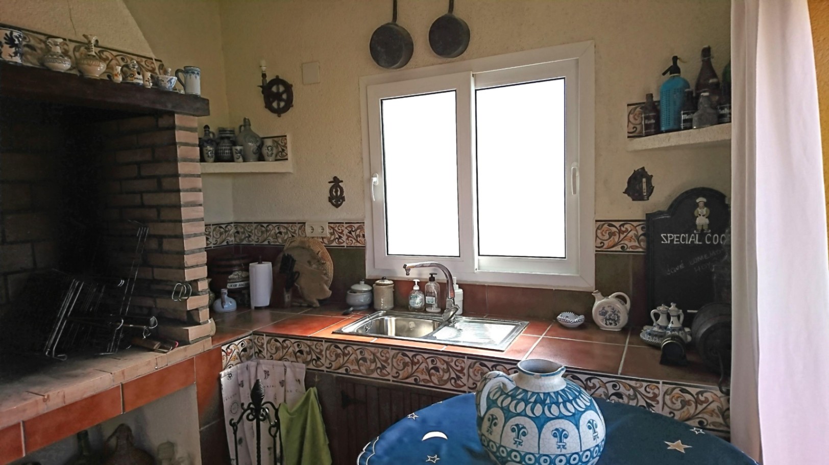 Rustic summer kitchen featuring a built-in brick barbecue, traditional tiled countertops, a stainless steel sink, and a white PVC window for natural light.