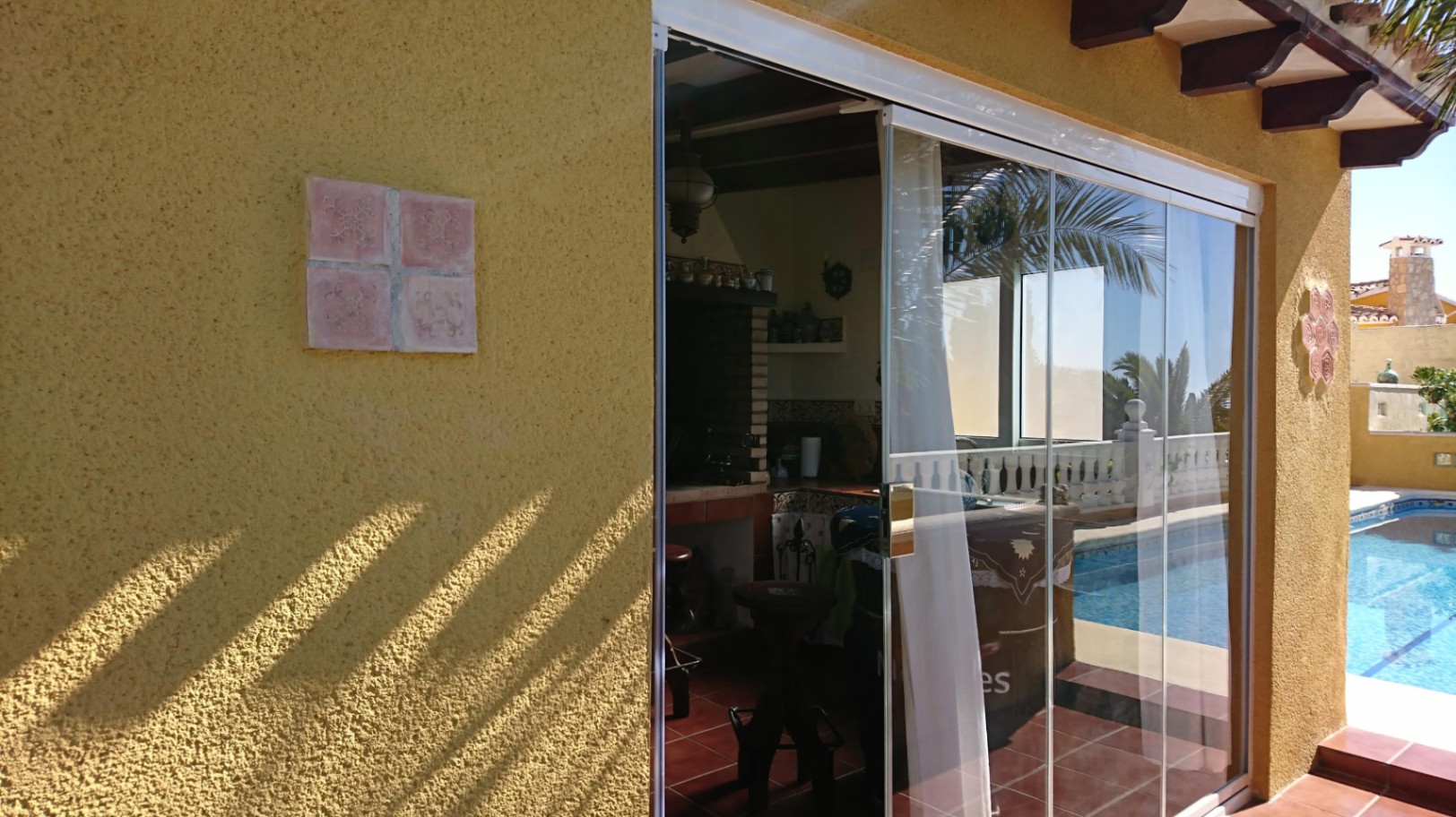 Summer kitchen enclosed with glass curtains, featuring a built-in brick barbecue, wooden ceiling beams, and direct access to the pool area.