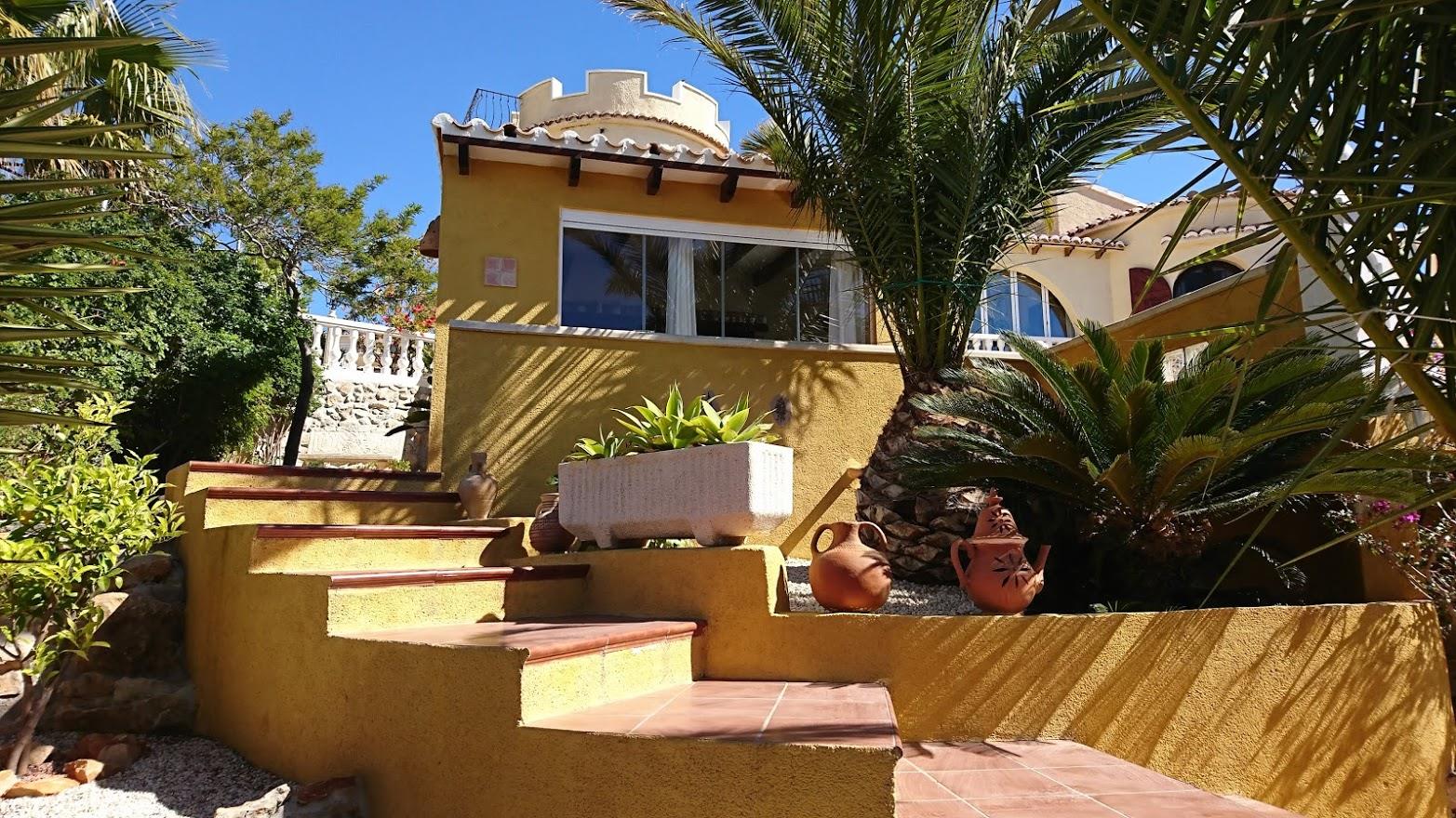 Terracotta exterior stairs lead to a Mediterranean villa featuring a distinctive crenellated tower, ochre walls, and landscaped gardens with palm trees.