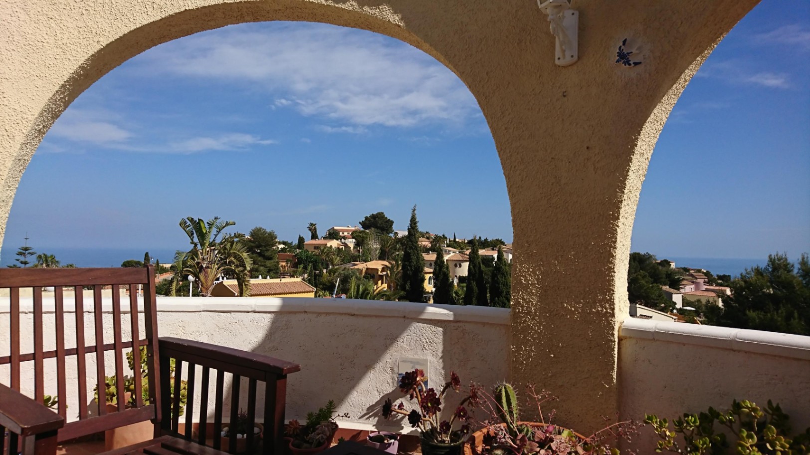 View from the arched terrace overlooking the Mediterranean Sea and surrounding greenery. The space features wooden seating, potted succulents, and traditional textured architecture.