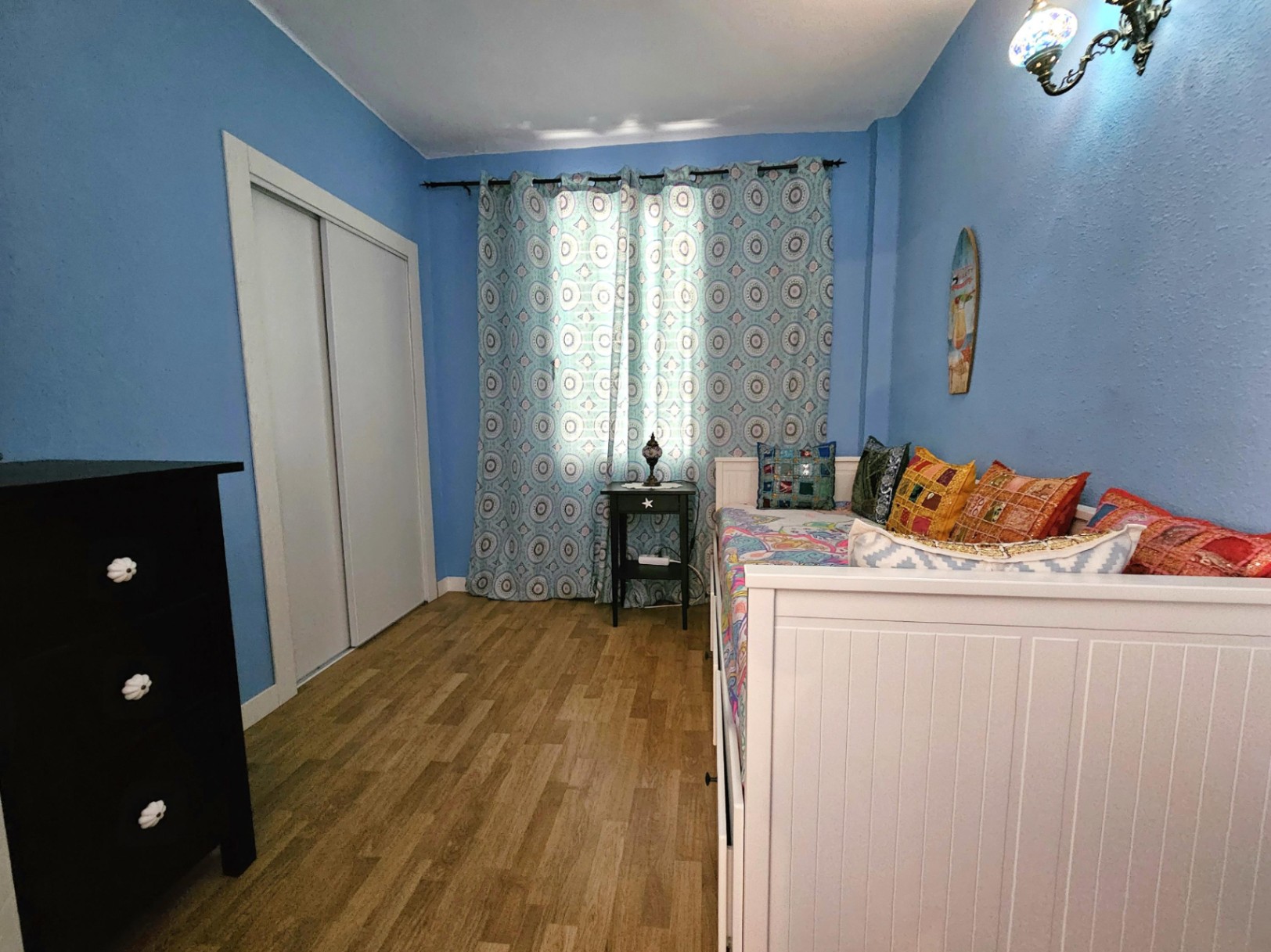 Bedroom with light blue walls, parquet flooring, and a white built-in wardrobe. It features a white daybed with colorful cushions and a dark chest of drawers.