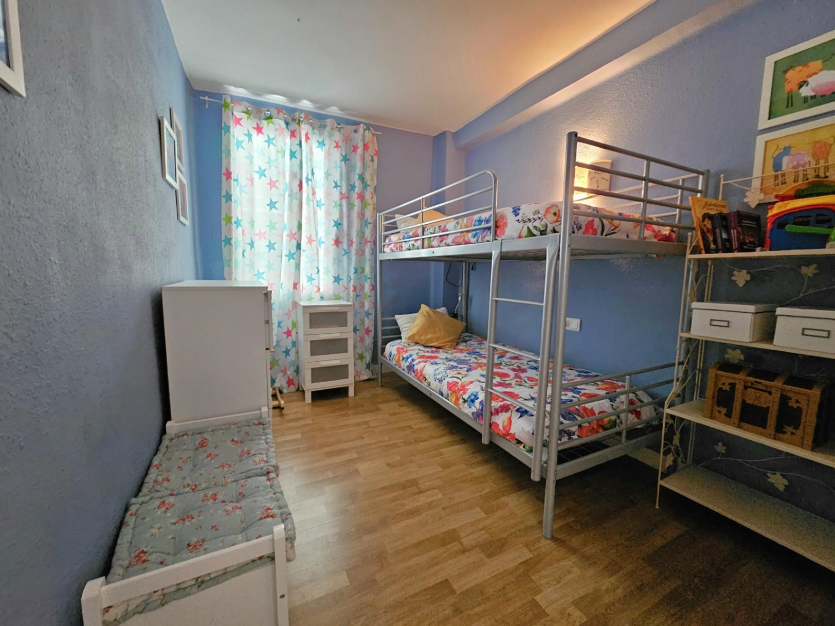 Children's bedroom featuring silver metal bunk beds, warm parquet flooring, and blue walls. The space includes white storage units and natural window light.