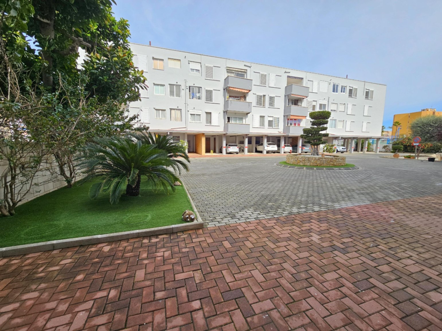 Exterior view of the residential complex featuring a paved courtyard, landscaped garden beds, and private covered parking bays.