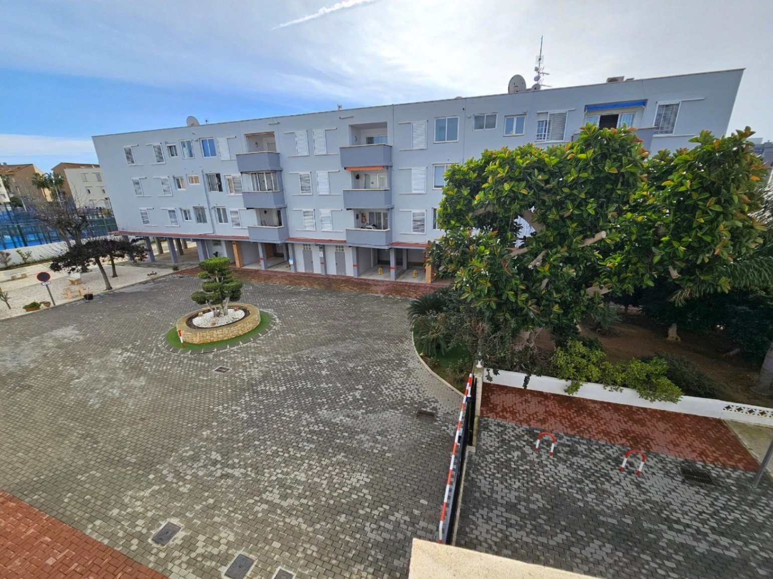 Exterior view of the residential complex featuring a paved courtyard, landscaped planters, and private covered parking bays beneath the building.
