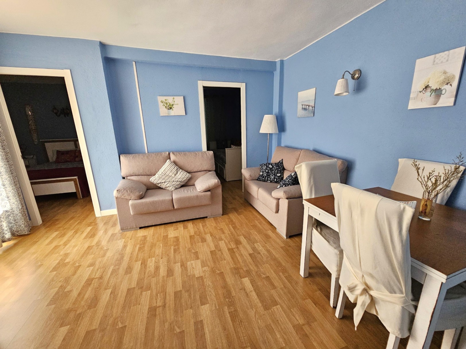 Living and dining area featuring blue walls, parquet flooring, and two beige sofas. The bright space connects directly to the adjacent bedrooms through white-framed doorways.