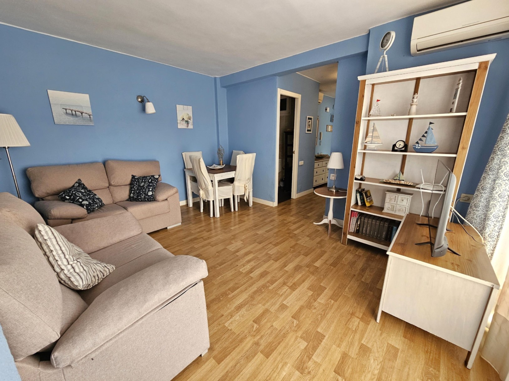 Living and dining space with blue walls, parquet flooring, and a beige sofa. Features include a wall-mounted AC unit and a white shelving unit with nautical decor.