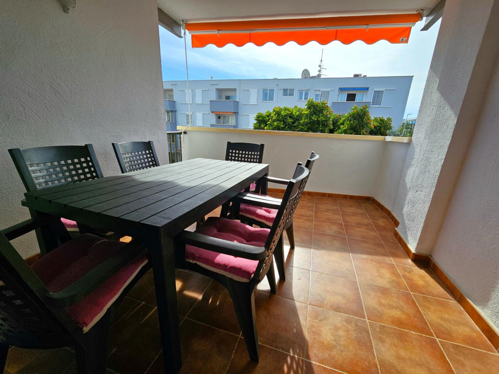 Outdoor terrace featuring terracotta floor tiles, a dark dining set for six with pink cushions, and a retractable orange awning for shade.