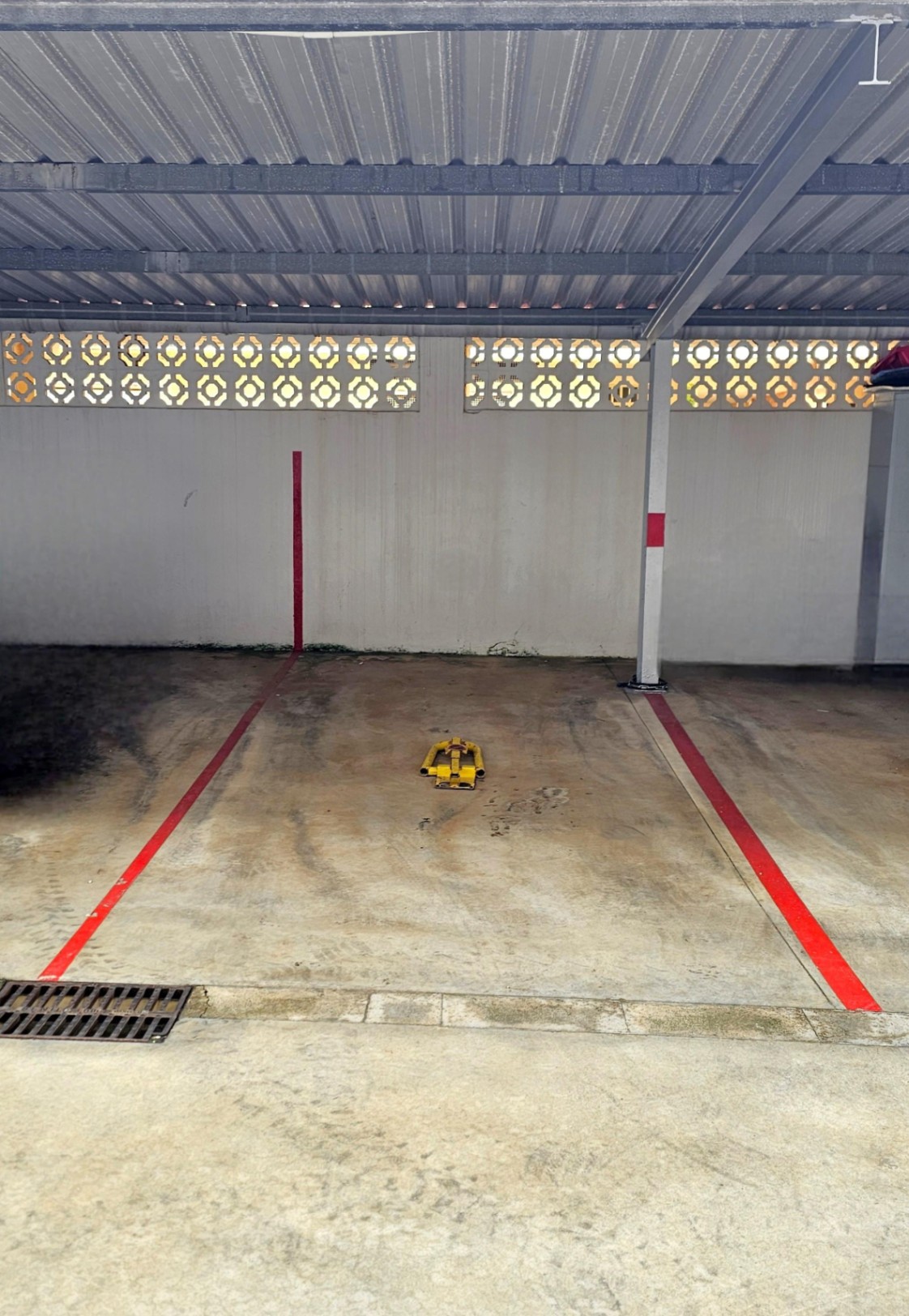 Private covered parking space featuring a corrugated metal roof, red boundary markings, and a yellow security bollard on a concrete surface.