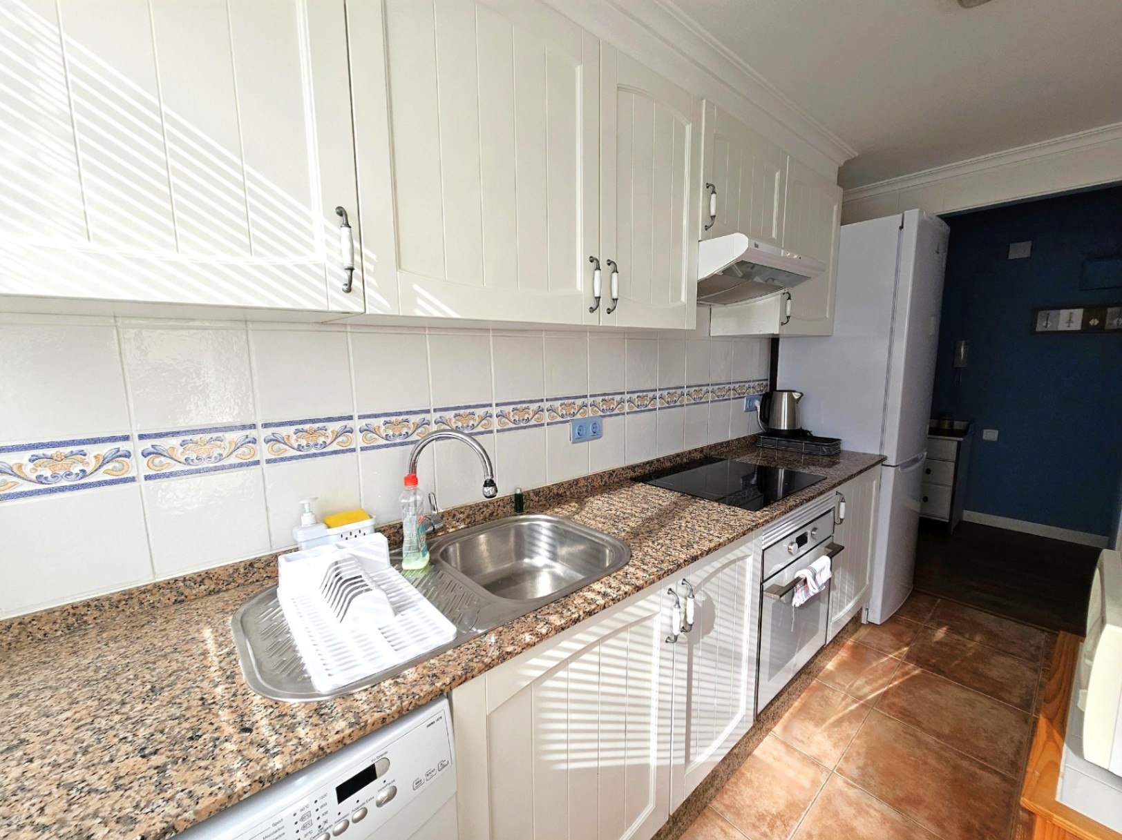 Renovated kitchen featuring white paneled cabinetry, granite countertops, and integrated appliances. The space includes a decorative tiled backsplash and terracotta flooring.