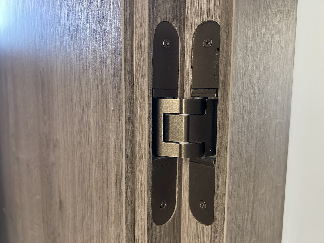 Close-up of a high-end concealed door hinge in a bronze finish, seamlessly integrated into the dark wood grain frame for a minimalist aesthetic.