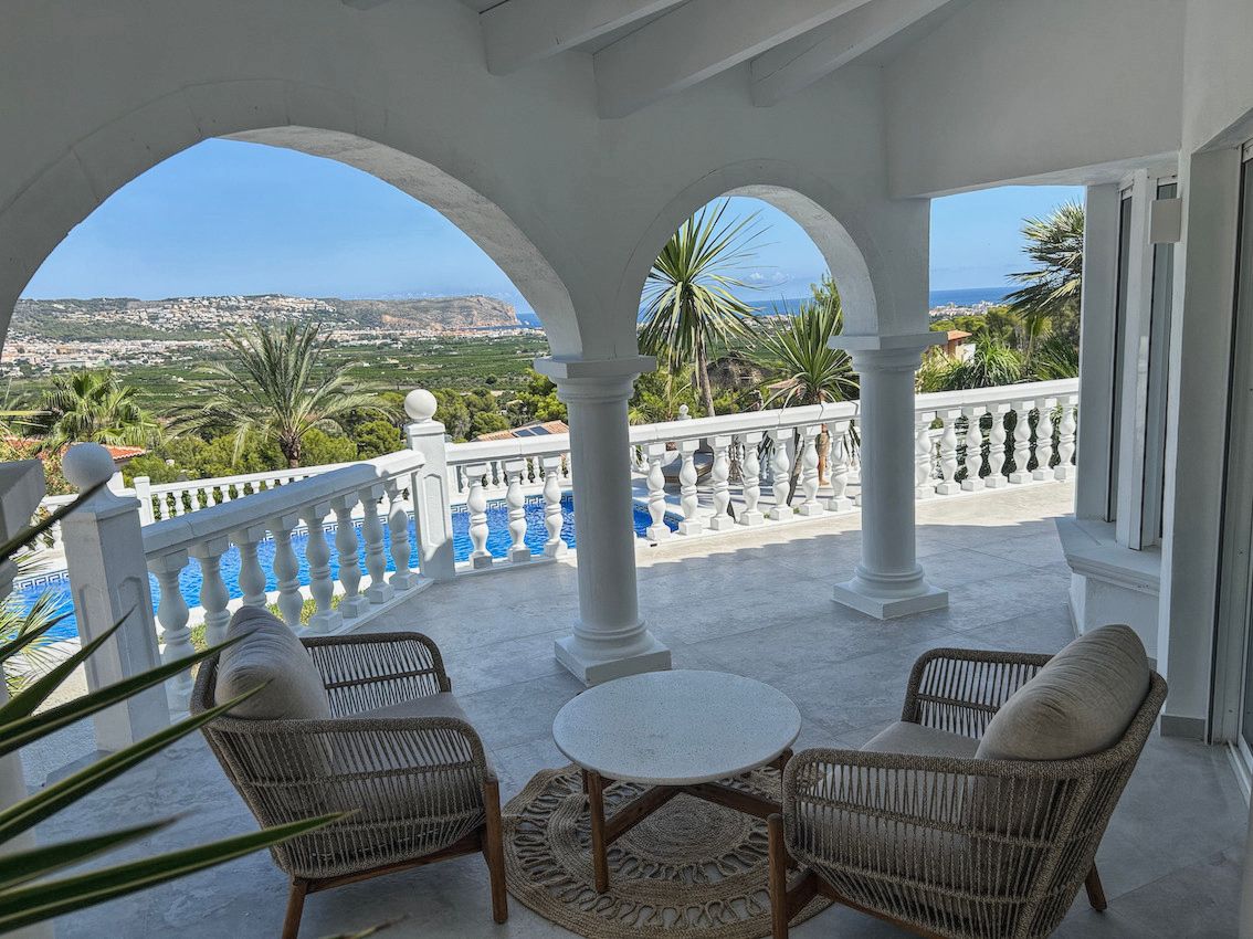 Covered terrace featuring white arches and columns, furnished with rattan armchairs. It overlooks the private pool with panoramic valley and sea views.