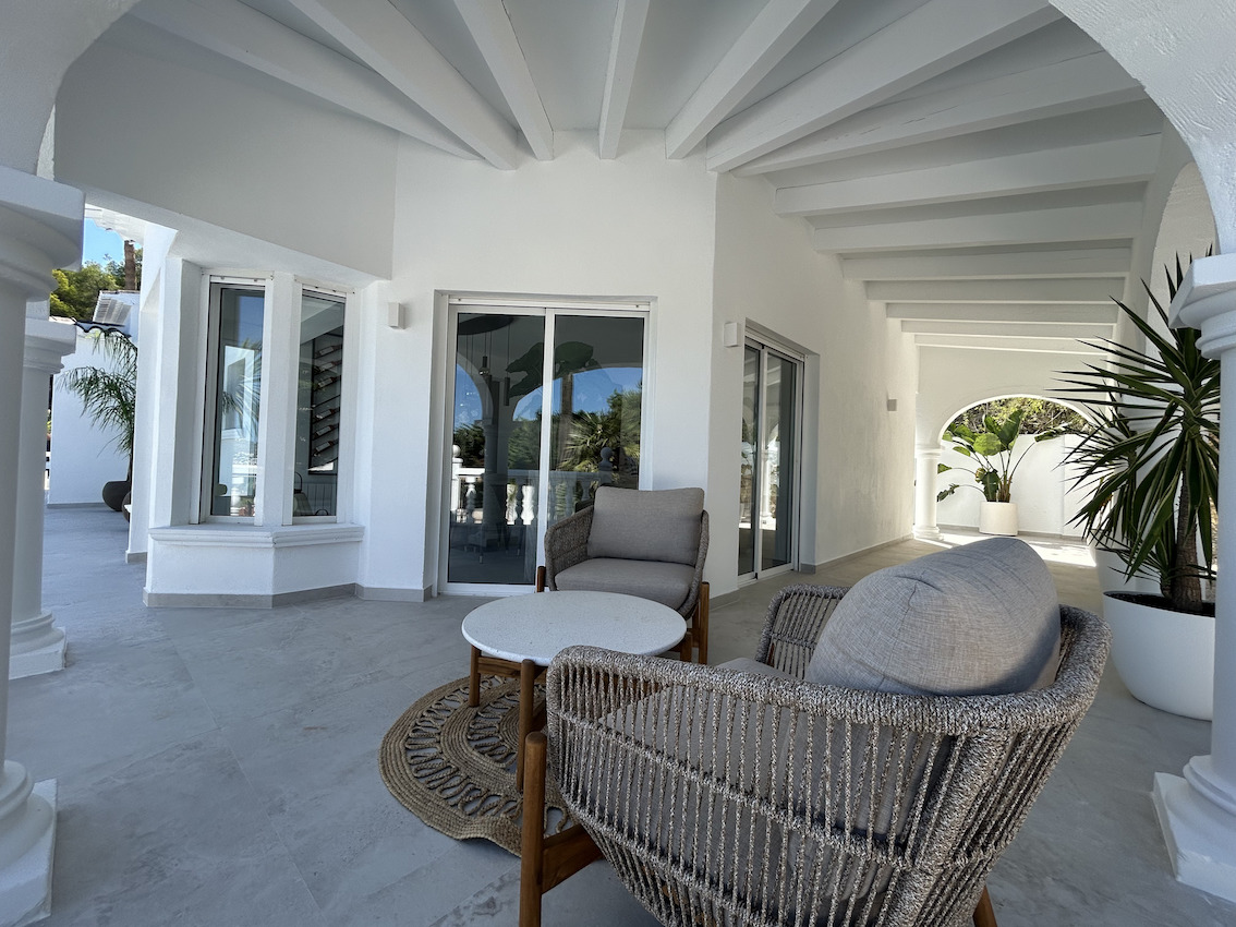 Covered terrace featuring white beamed ceilings, contemporary woven armchairs, and grey stone flooring, seamlessly connecting to the villa's interior.