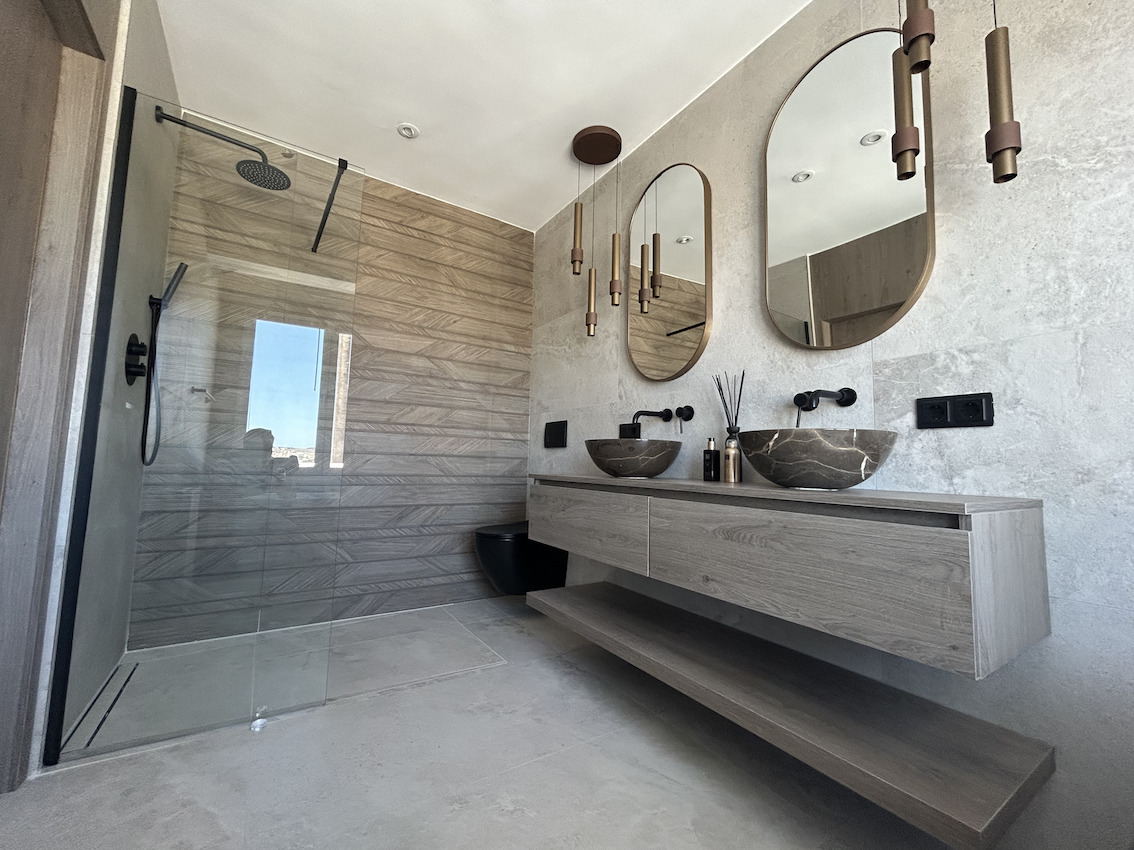 Modern bathroom featuring a double stone vanity, matte black fixtures, oval mirrors, and a walk-in shower with textured wood-effect tiling.