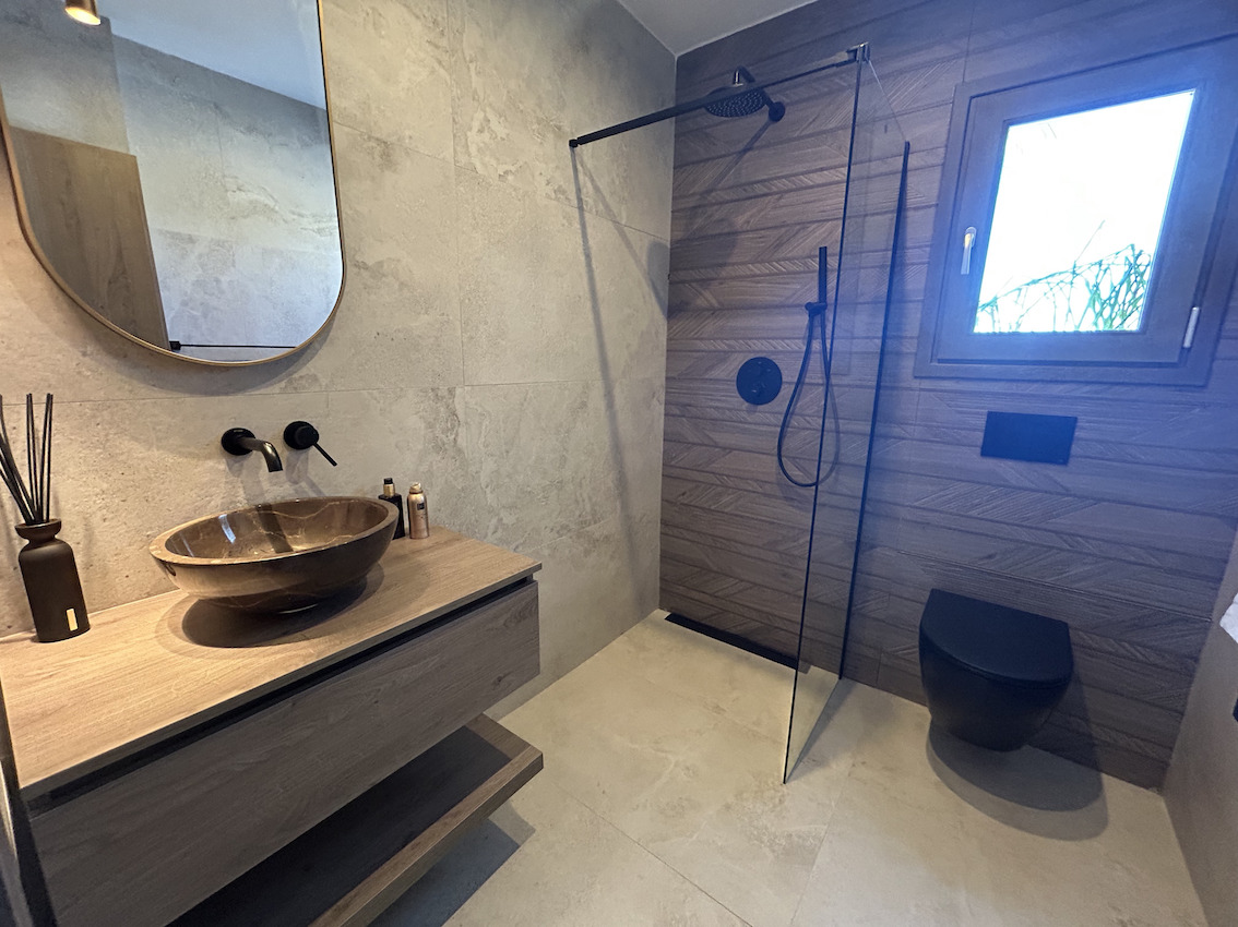 Modern bathroom featuring a walk-in shower with black fixtures, a stone vessel sink on a floating wooden vanity, and textured wall tiling.