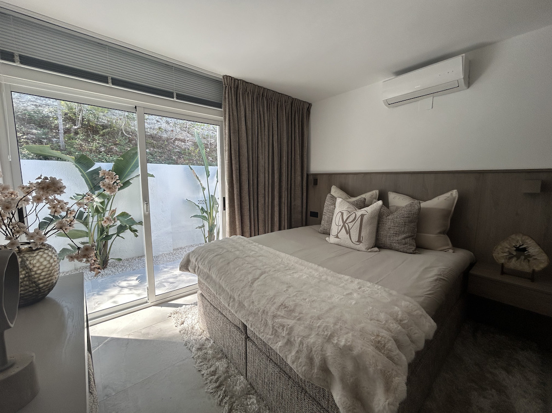 Modern bedroom with direct patio access via sliding glass doors, featuring split air conditioning and a custom wood-paneled headboard.