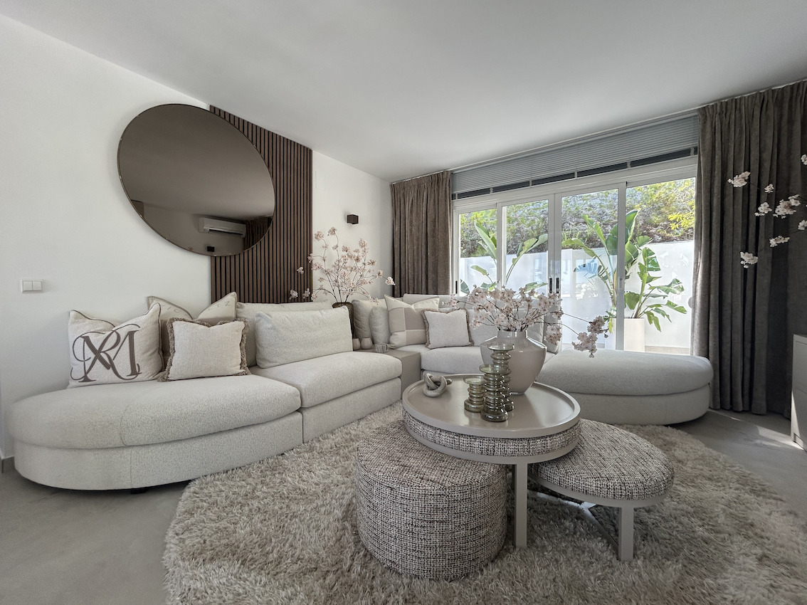 Modern living area featuring a curved modular sofa, wood-slat accent wall with a large circular mirror, and sliding glass doors opening to a lush terrace.