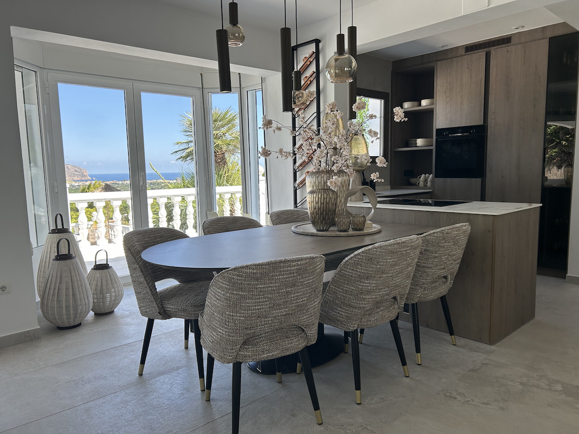 Open-concept dining area featuring an oval table and integrated designer kitchen with wood cabinetry, offering panoramic sea views through large windows.
