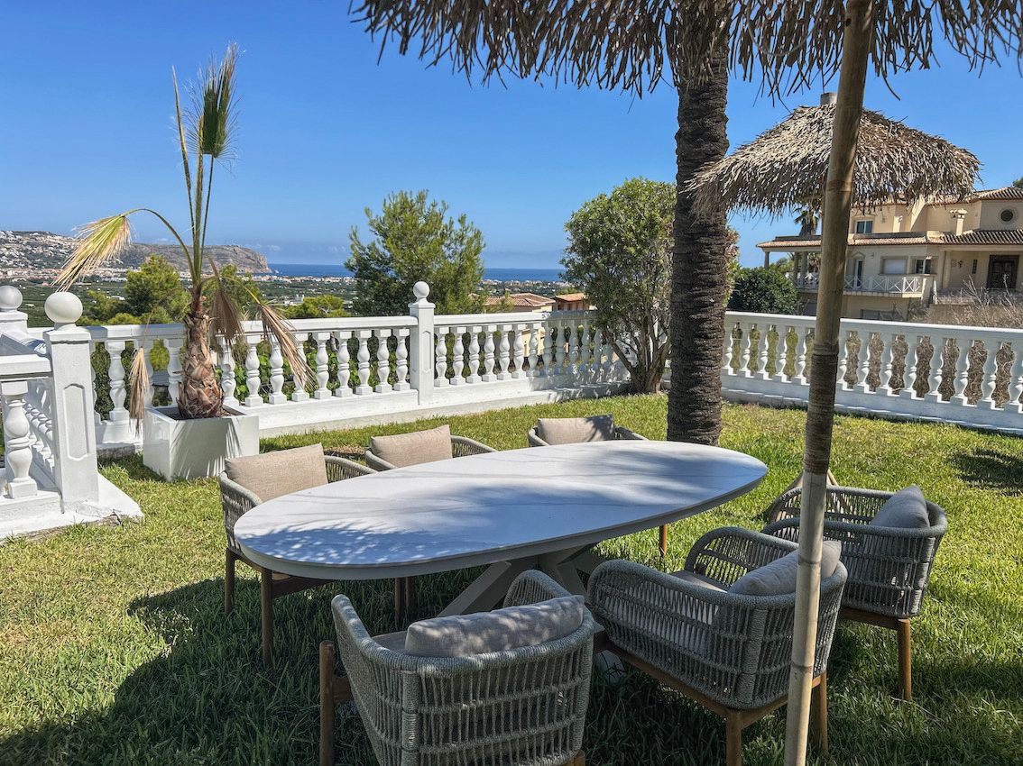 Outdoor dining area on a natural lawn featuring an oval table, woven chairs, and a white balustrade overlooking the valley and sea.