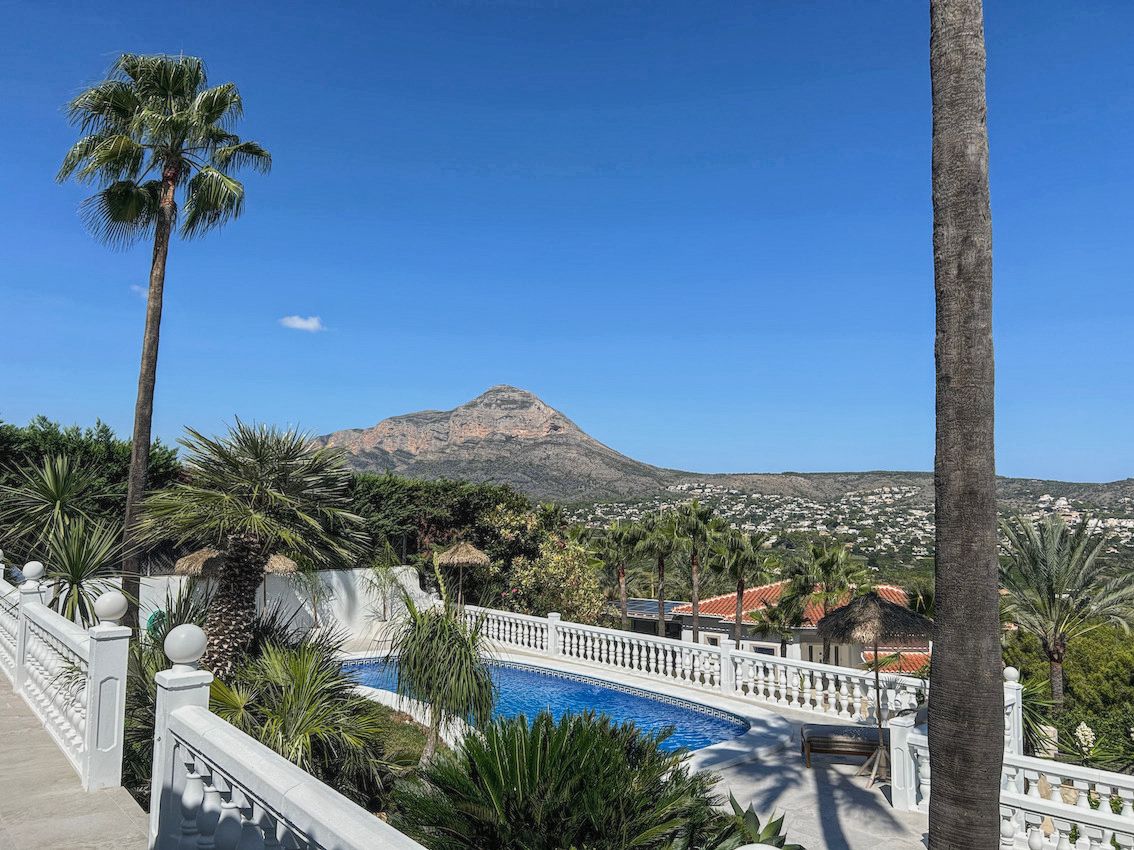 Outdoor swimming pool area featuring white balustrades, tropical palms, and clear panoramic views of the mountain and surrounding valley.