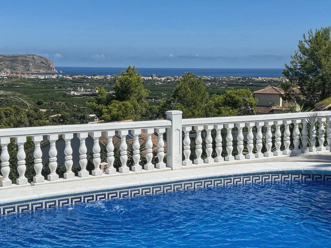 Swimming pool with mosaic tiling and a white stone balustrade overlooking the Mediterranean Sea, coastal valley, and distant cliffs.
