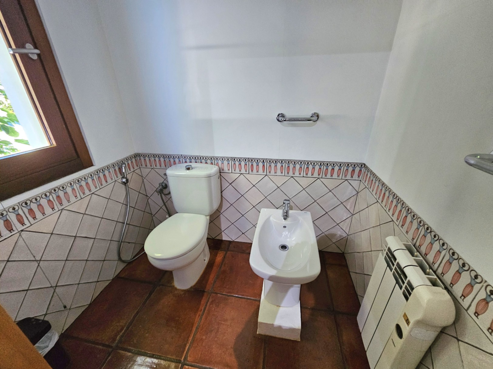 Bathroom area featuring a toilet and bidet, set against handmade terracotta flooring and walls with unique hand-painted ceramic borders.
