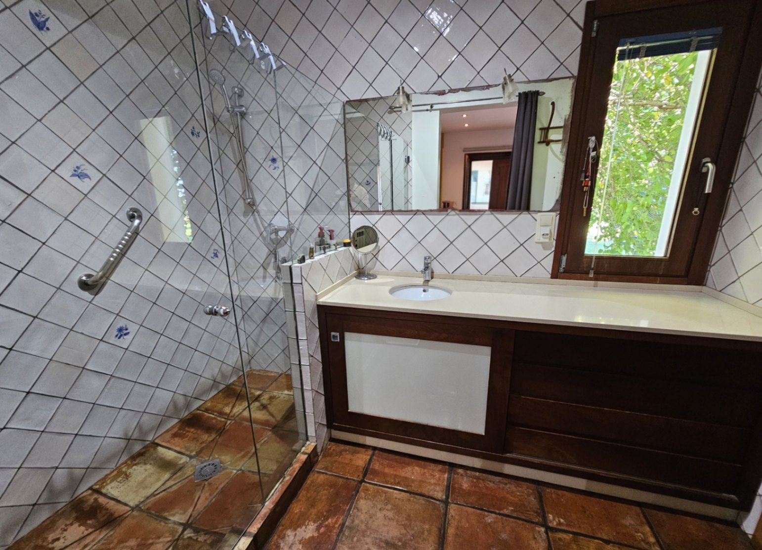 Bathroom featuring hand-painted wall tiles, a walk-in glass shower, and handmade clay flooring. A wooden vanity with a white countertop sits beside a window with garden views.