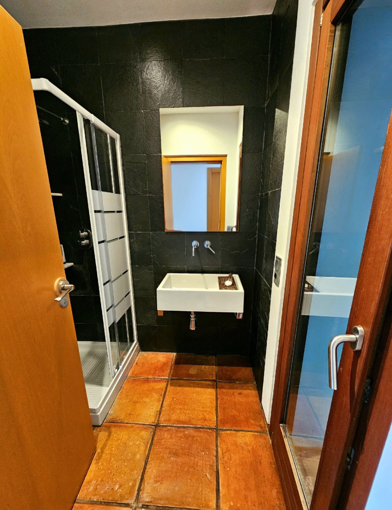 Bathroom featuring handmade terracotta floor tiles, dark textured stone walls, a wall-mounted rectangular basin, and a glass shower enclosure.