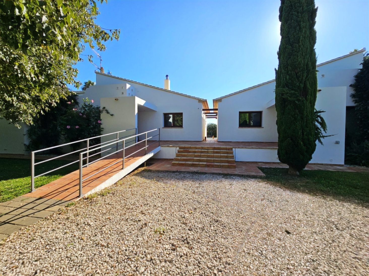 Exterior view of the villa's two wings featuring a terracotta terrace, a metal accessibility ramp, and a gravel driveway bordered by Mediterranean greenery.