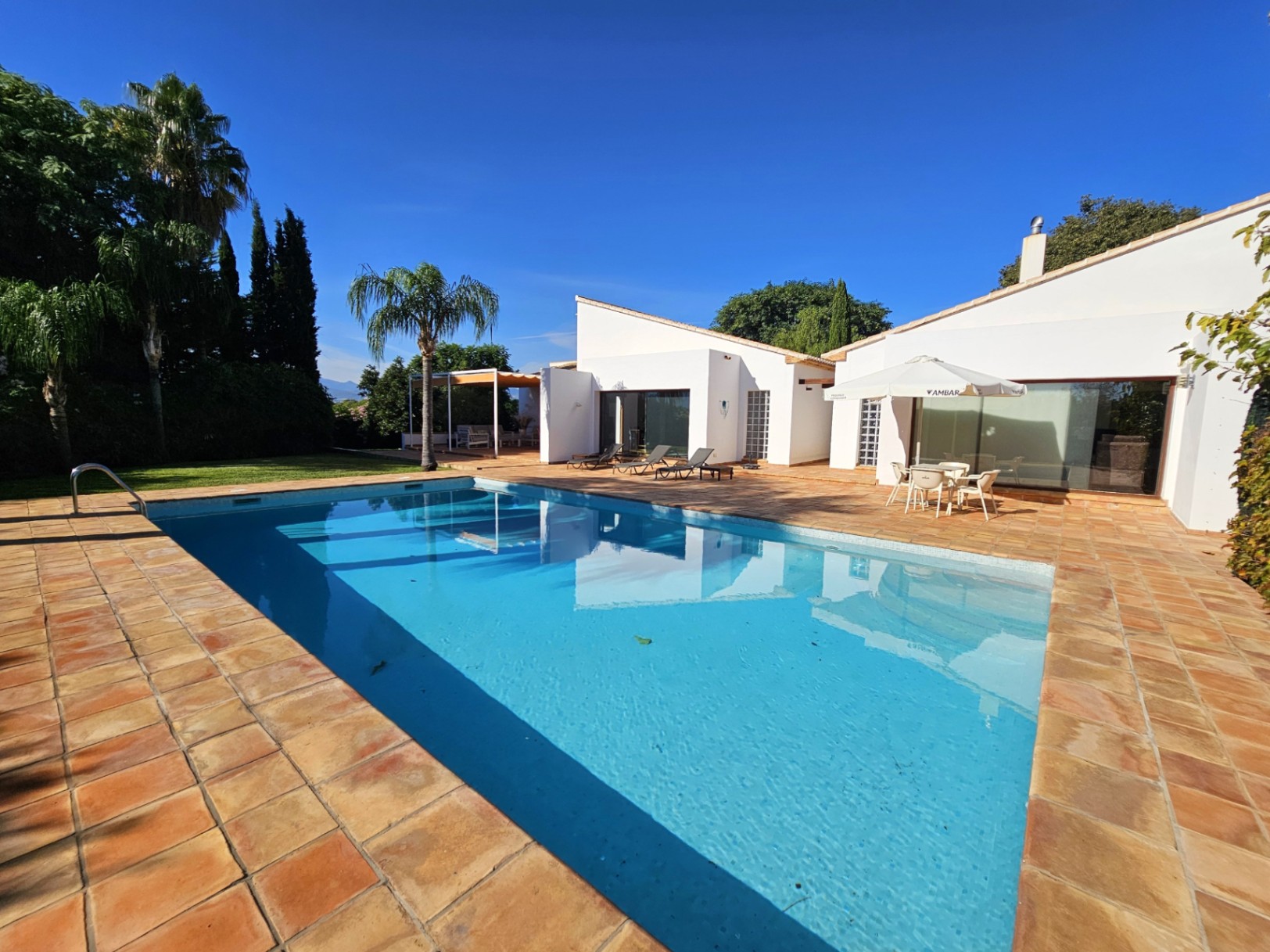 Large outdoor swimming pool set within a terracotta tiled terrace, featuring reflections of the white villa architecture and lush Mediterranean landscaping.