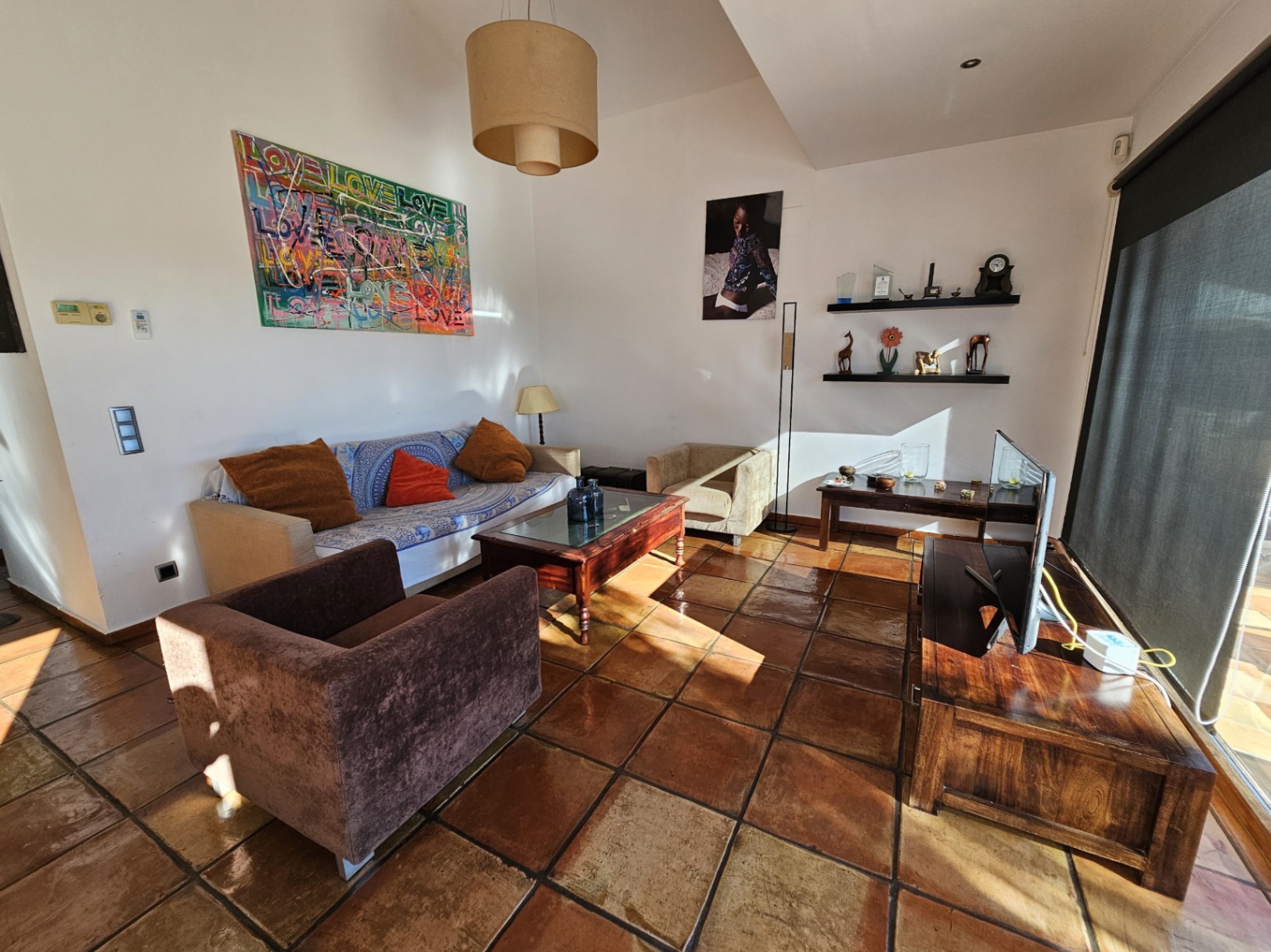 Living area featuring handmade terracotta tile flooring, wooden furniture, and vibrant artwork. Natural light streams through large windows equipped with security glass.