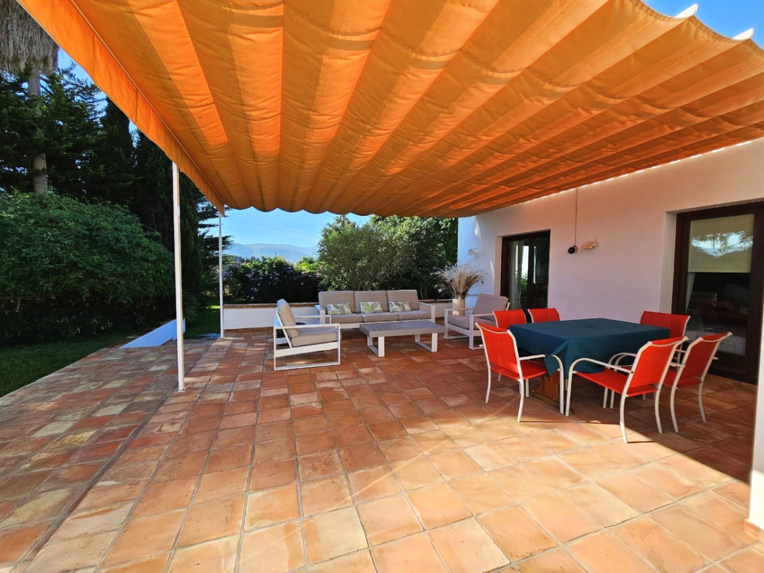 Outdoor terrace featuring handmade clay tiles and a large orange retractable awning. It includes a dining area and lounge space overlooking the lush garden.