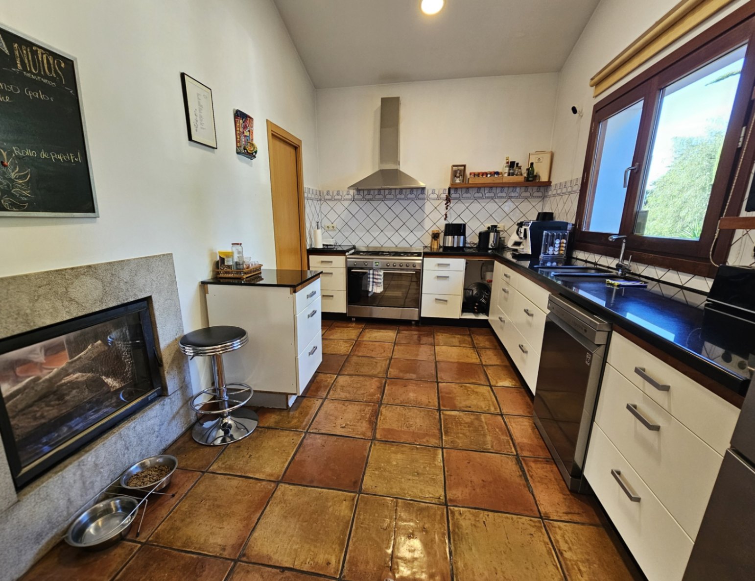 Rustic kitchen featuring handmade clay tile flooring, dark countertops, and a double-sided fireplace integrated into a natural stone wall.