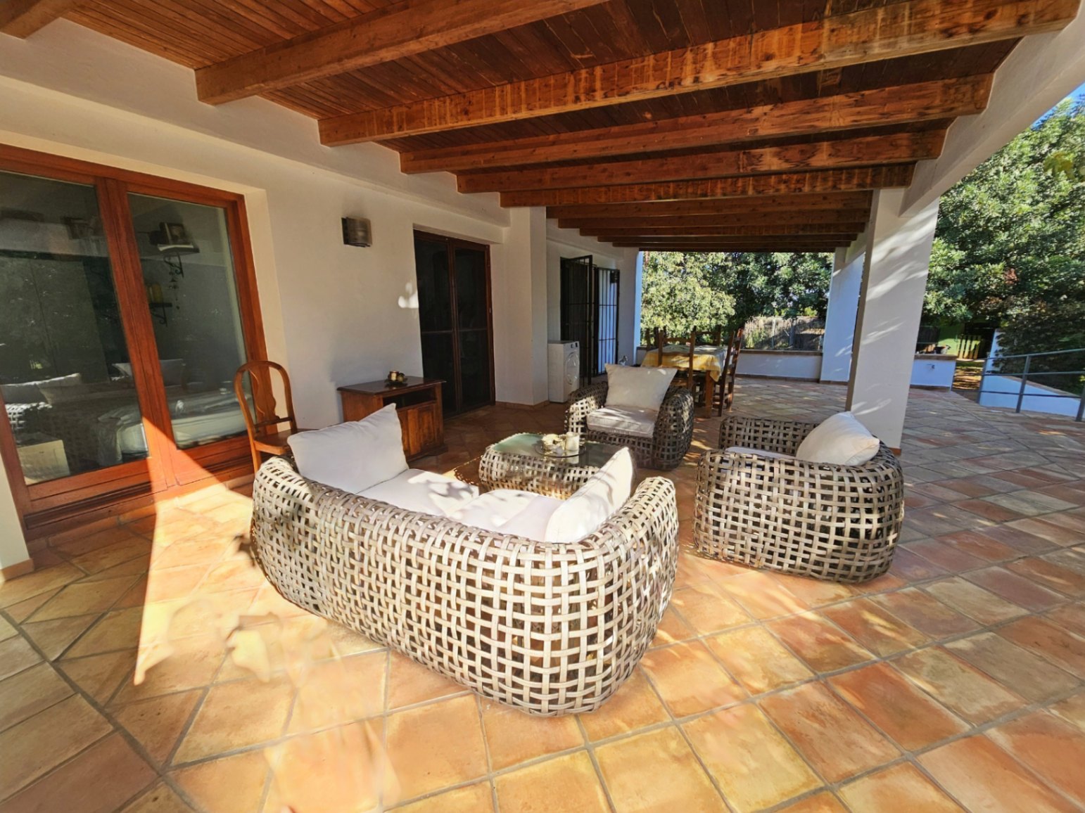 Spacious covered terrace featuring exposed wooden beams, handmade terracotta flooring, and a woven rattan lounge set overlooking the garden.