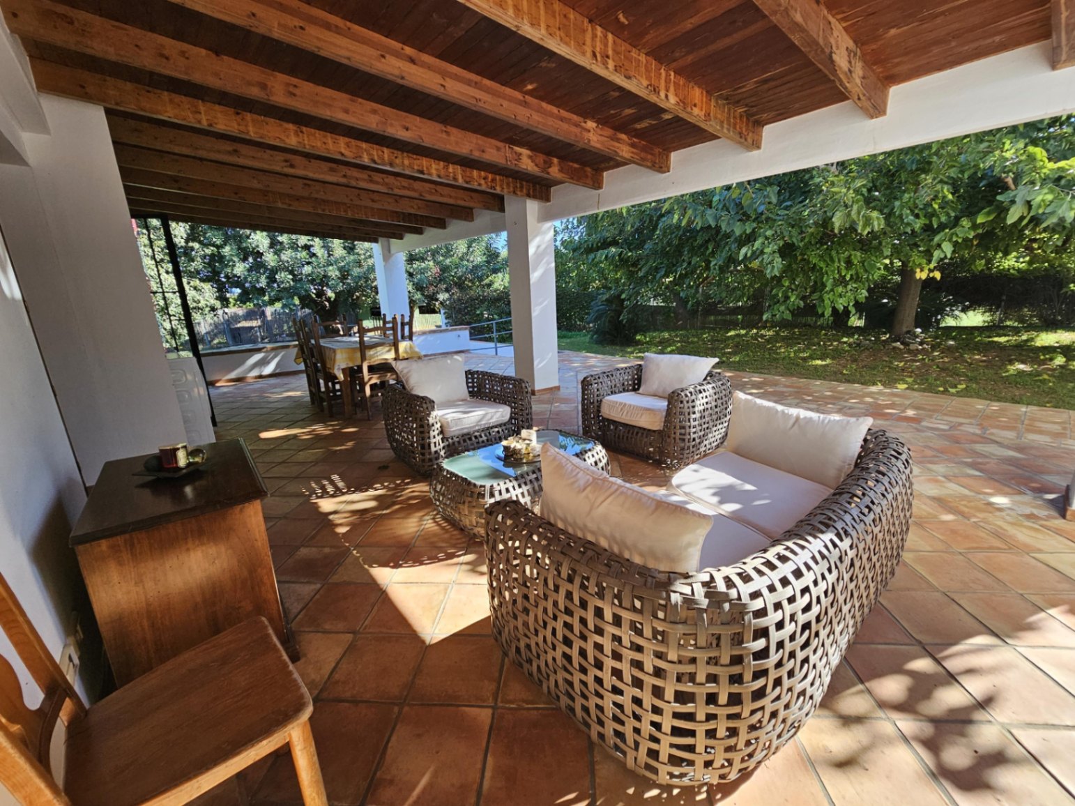 Spacious covered terrace featuring exposed wooden beams, handmade terracotta flooring, and woven wicker lounge furniture overlooking the garden.