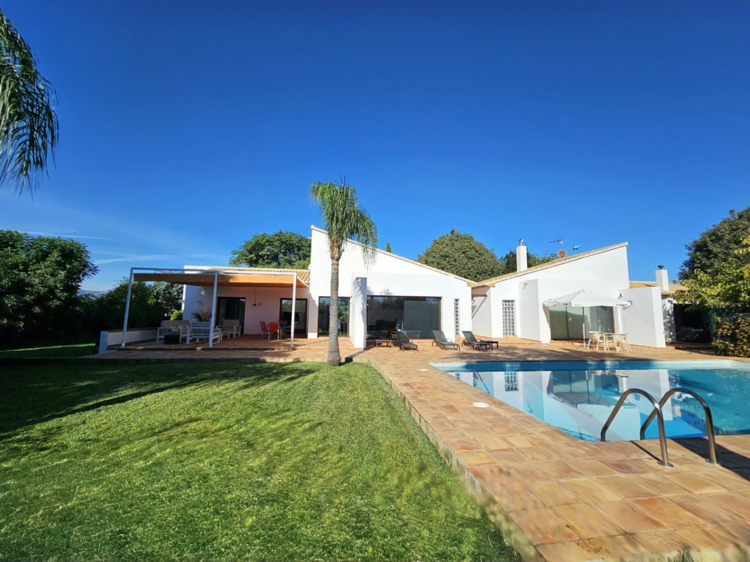 Villa exterior featuring a swimming pool with handmade clay tile decking, a manicured lawn, and a covered terrace integrated into the modern white architecture.