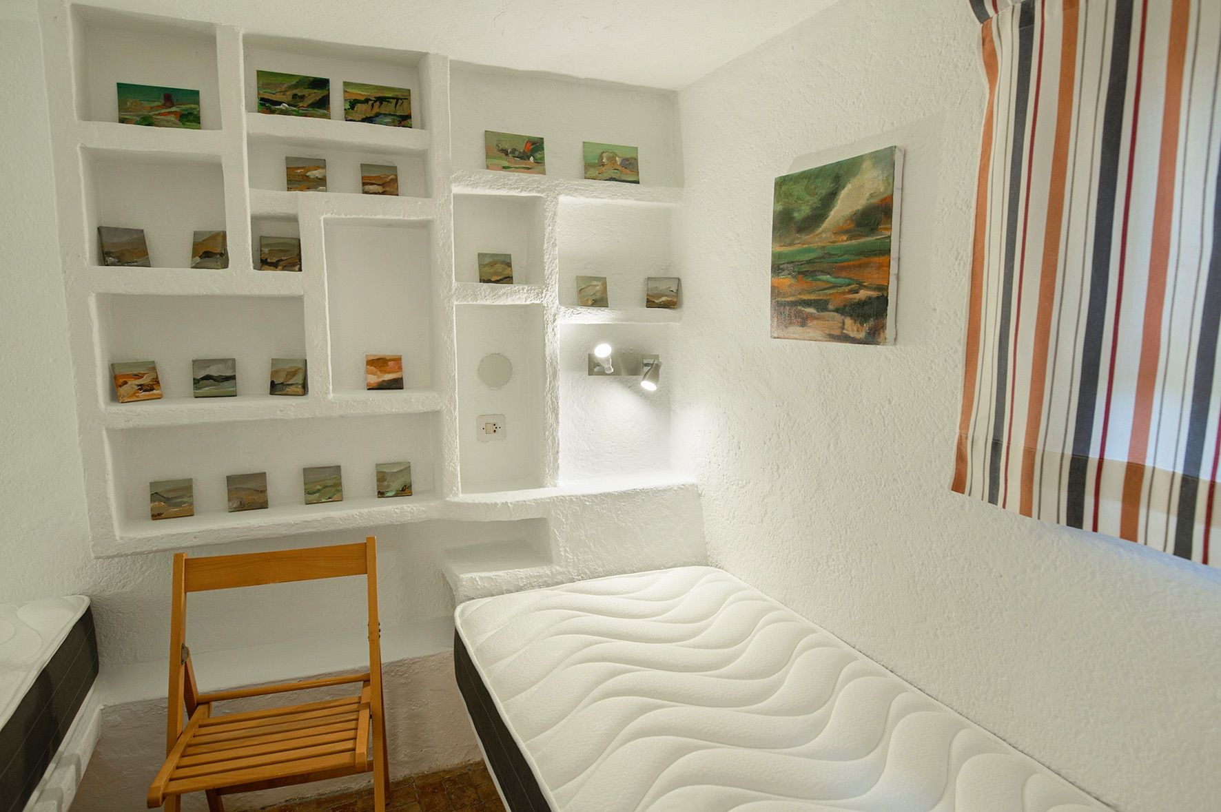 Bedroom featuring white textured masonry walls with built-in shelving niches for artwork, a single mattress, and integrated spotlighting.
