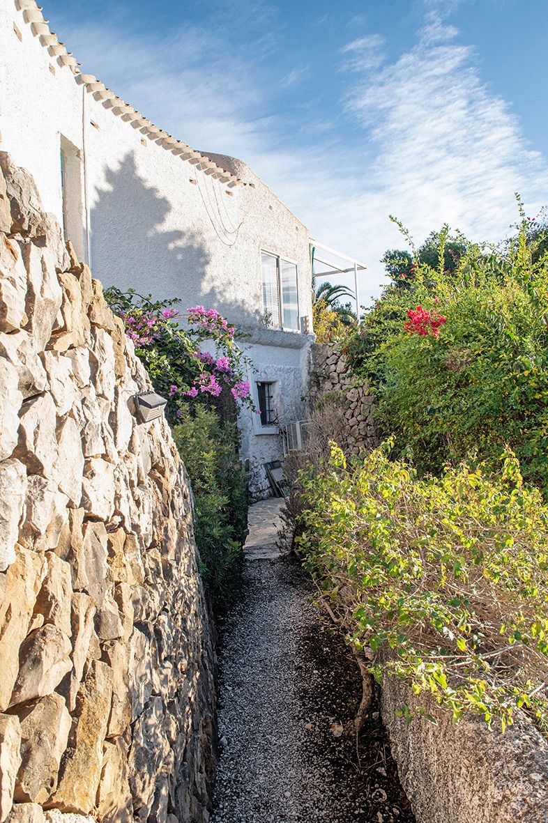 Exterior garden path bordered by traditional dry stone walls and lush Mediterranean plants, leading alongside the white-washed villa facade.