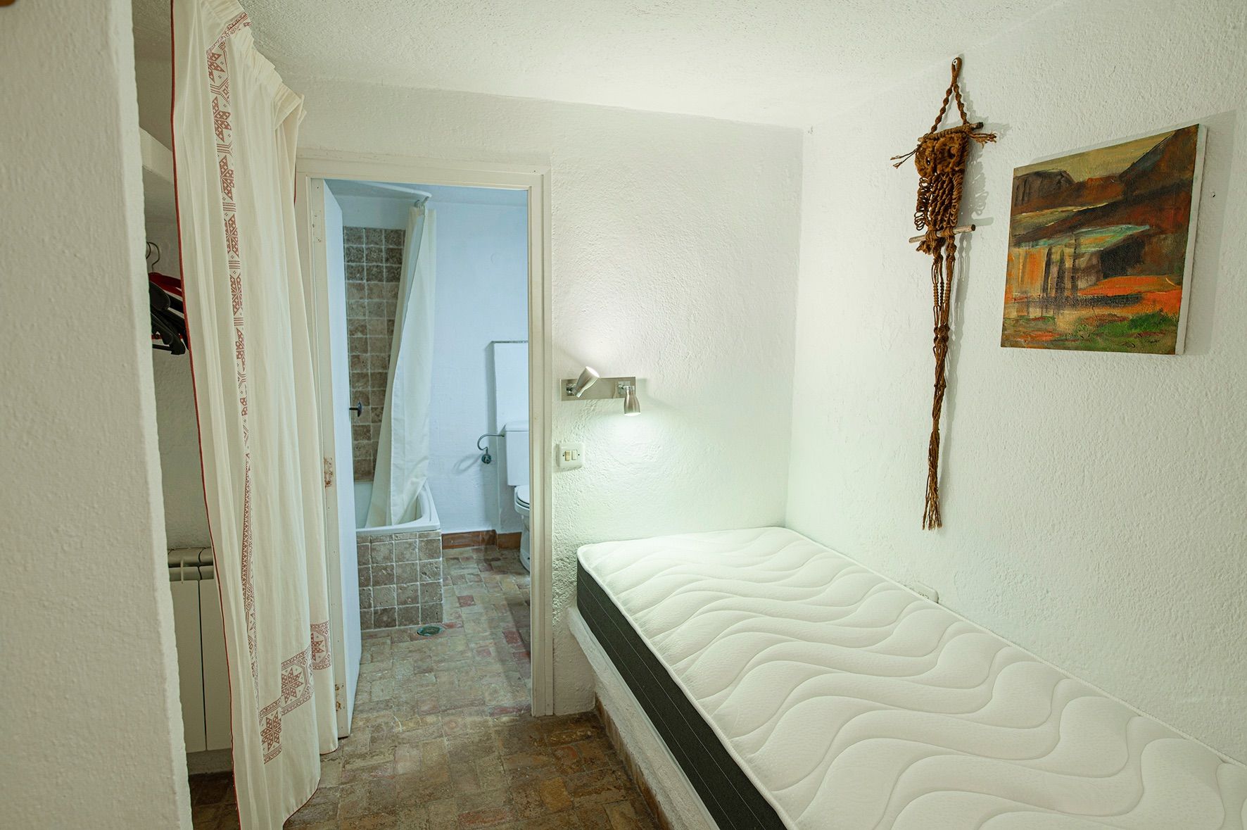 Functional bedroom featuring a single bed on a built-in base, textured white walls, and an ensuite bathroom with earth-toned rustic tiling.