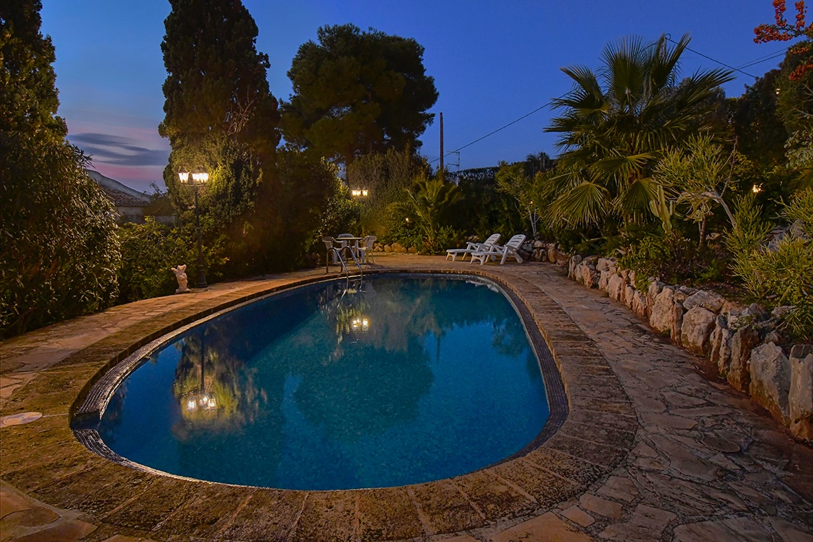 Illuminated swimming pool at dusk, featuring a natural stone terrace and a mature Mediterranean garden with palms and cypress trees.