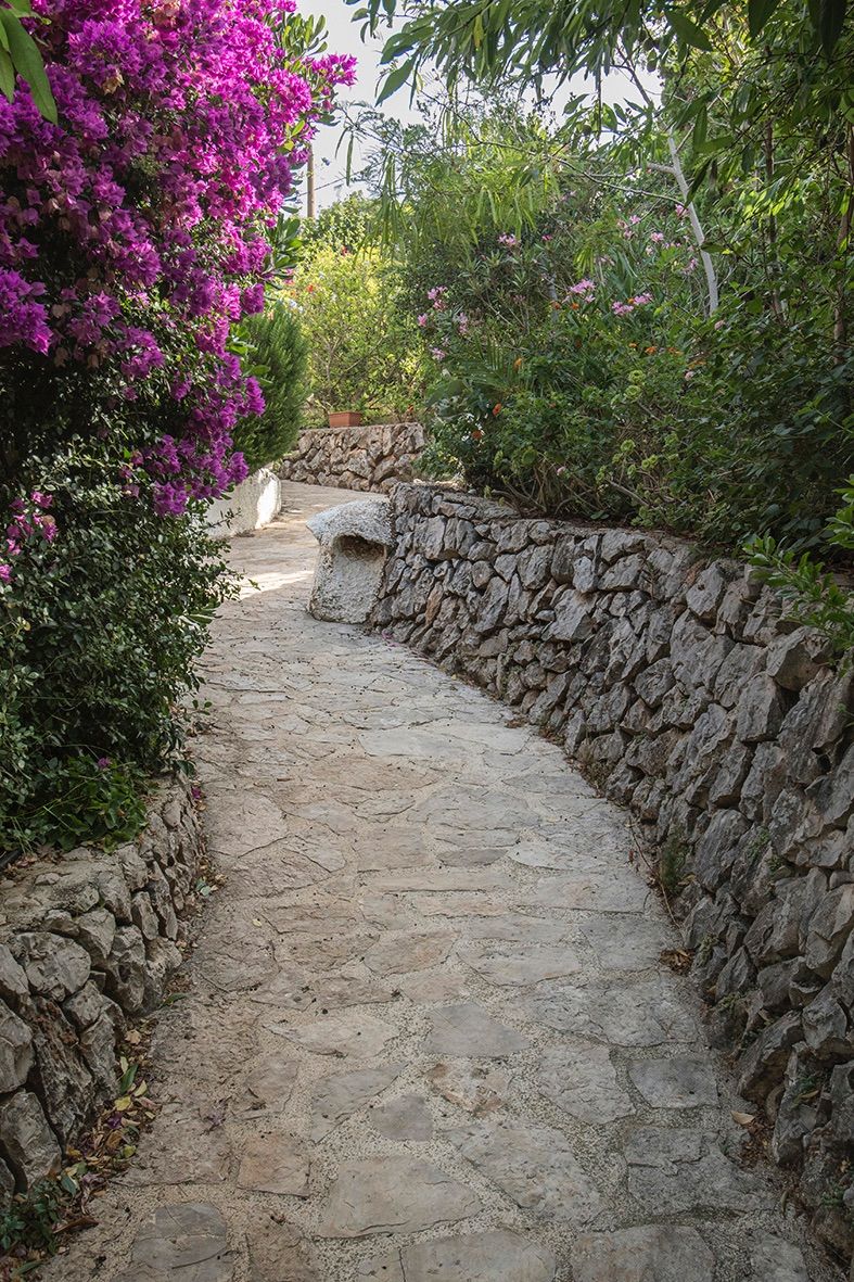 Irregular stone-paved garden path bordered by traditional dry stone walls and lush Mediterranean greenery, including vibrant bougainvillea.